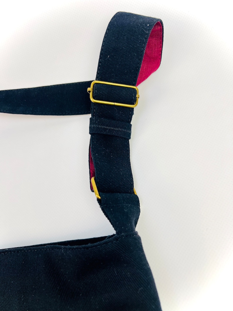 K&ouml;nnte beinhalten: Eine schwarze Canvas-Tragetasche mit einem schwarzen und roten Riemen und einer goldenen Schnalle. Der Riemen ist verstellbar.