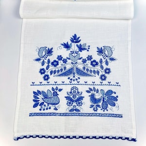 Peut inclure: Chemin de table en lin blanc avec broderie florale et d'oiseaux bleue. Le motif pr&eacute;sente des fleurs, des feuilles et des oiseaux stylis&eacute;s, avec une bordure bleue festonn&eacute;e. Une pi&egrave;ce d&eacute;corative pour une table.