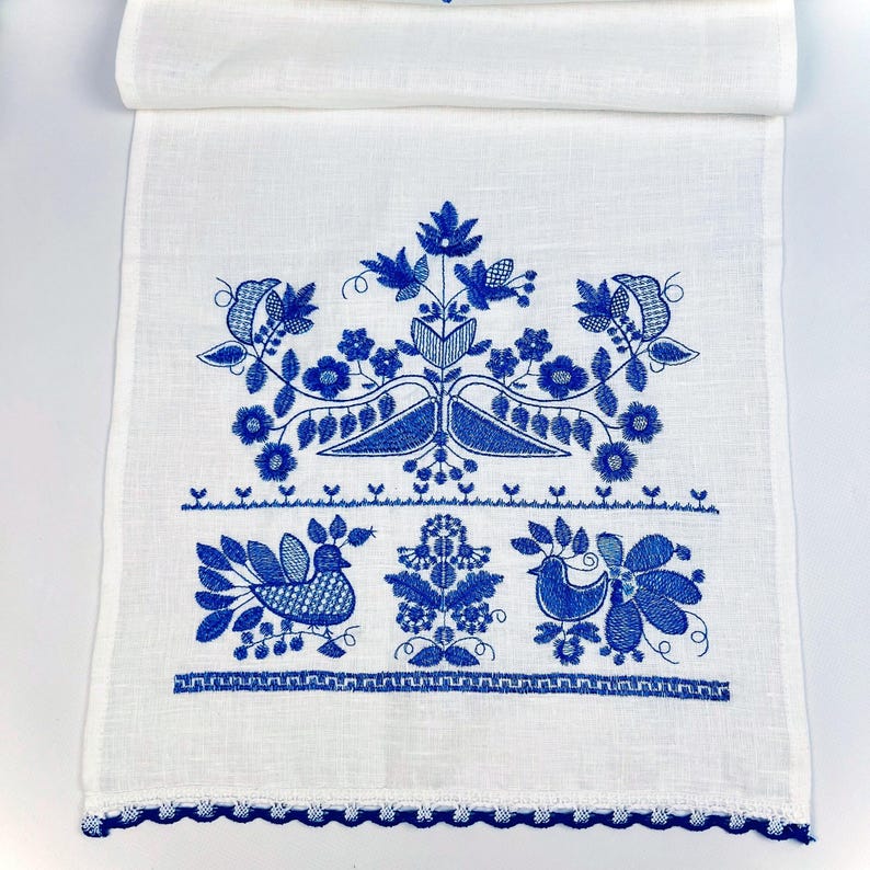 Peut inclure: Chemin de table en lin blanc avec broderie florale et d'oiseaux bleue. Le motif pr&eacute;sente des fleurs, des feuilles et des oiseaux stylis&eacute;s, avec une bordure bleue festonn&eacute;e. Une pi&egrave;ce d&eacute;corative pour une table.