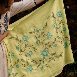 May include: A yellow linen scarf with a floral embroidery design. The flowers are a light blue color with dark green stems and leaves.