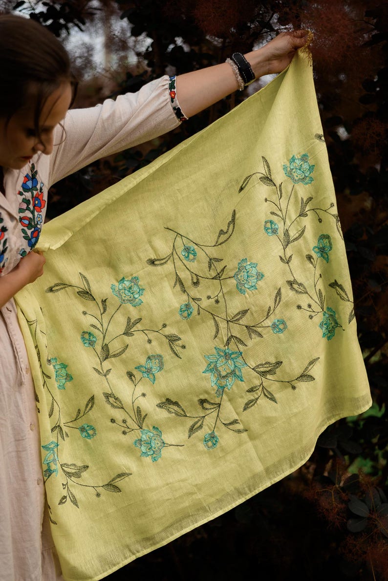 May include: A yellow linen scarf with a floral embroidery design. The flowers are a light blue color with dark green stems and leaves.