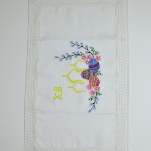 May include: White linen Easter table runner with embroidered Easter eggs, flowers, and branches. The runner has a crocheted lace border. The letters XB are embroidered in yellow. A decorative piece for the Easter holiday.