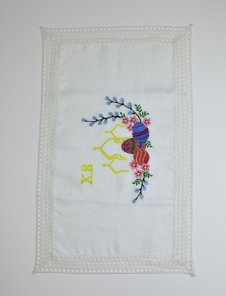 May include: White linen Easter table runner with embroidered Easter eggs, flowers, and branches. The runner has a crocheted lace border. The letters XB are embroidered in yellow. A decorative piece for the Easter holiday.