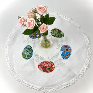 以下が含まれることがあります： レースの縁取りと刺繍された花の模様が施された白いリネン製のテーブルクロス。ピンクのバラの花瓶の周りに、カラフルな布製のイースターエッグが5つ並んでいます。