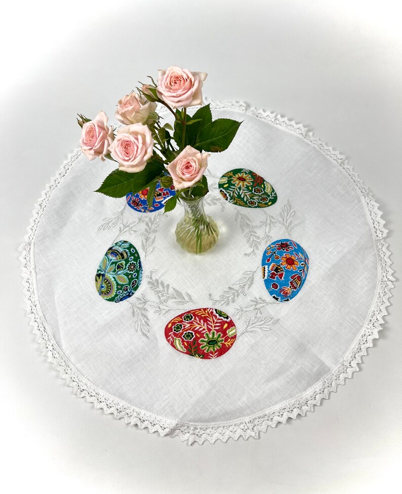 以下が含まれることがあります： レースの縁取りと刺繍された花の模様が施された白いリネン製のテーブルクロス。ピンクのバラの花瓶の周りに、カラフルな布製のイースターエッグが5つ並んでいます。