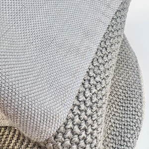 K&ouml;nnte beinhalten: Nahaufnahme einer gefalteten, grauen Strickdecke. Die Decke hat ein strukturiertes Design mit unterschiedlichen Strickmustern. Die Farbpalette besteht aus verschiedenen Graut&ouml;nen, die eine weiche und gem&uuml;tliche &Auml;sthetik erzeugen. Die Decke scheint aus einem weichen, warmen Material zu bestehen.