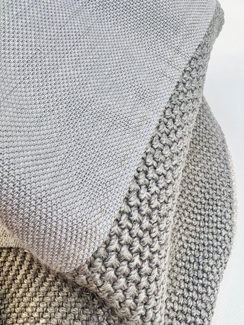 K&ouml;nnte beinhalten: Nahaufnahme einer gefalteten, grauen Strickdecke. Die Decke hat ein strukturiertes Design mit unterschiedlichen Strickmustern. Die Farbpalette besteht aus verschiedenen Graut&ouml;nen, die eine weiche und gem&uuml;tliche &Auml;sthetik erzeugen. Die Decke scheint aus einem weichen, warmen Material zu bestehen.