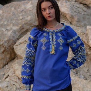 May include: A blue embroidered blouse with a geometric pattern in yellow and white thread. The blouse has long sleeves and a round neckline.