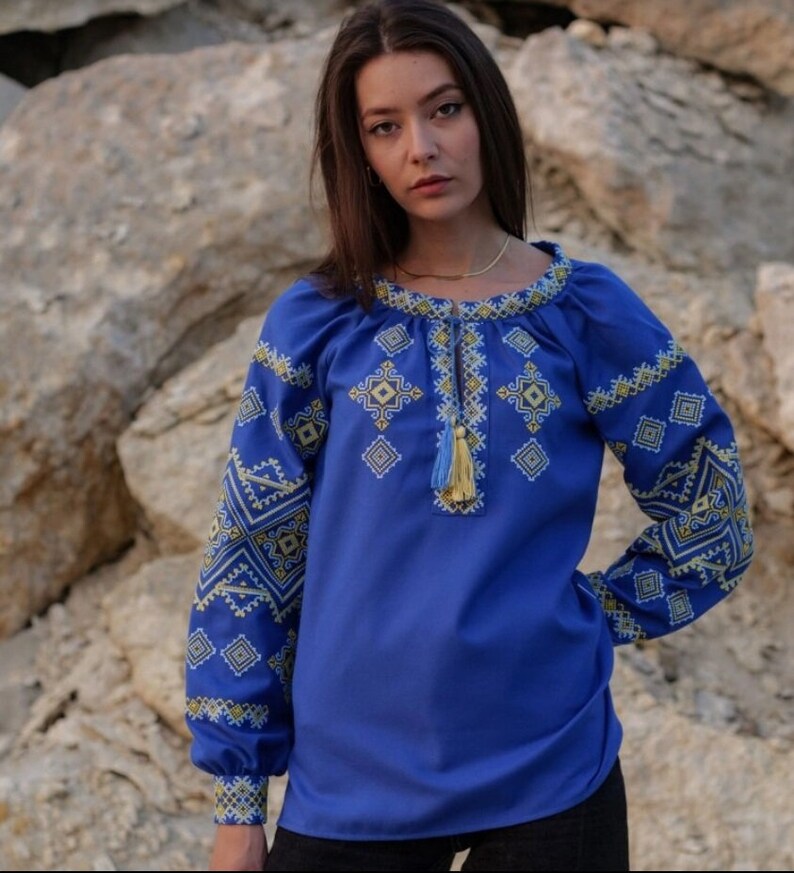 May include: A blue embroidered blouse with a geometric pattern in yellow and white thread. The blouse has long sleeves and a round neckline.