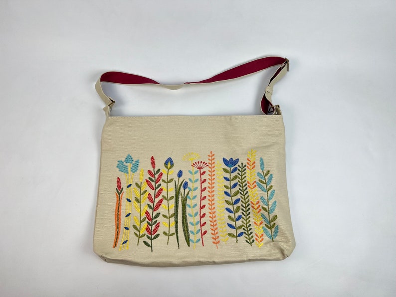 K&ouml;nnte beinhalten: Eine beige Canvas-Tragetasche mit einem roten Schultergurt und einem gestickten Design aus bunten Blumen.