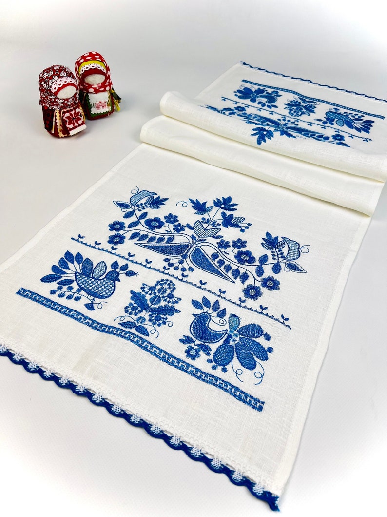 Peut inclure: Chemin de table en lin blanc avec broderie florale et d'oiseaux bleus. Le chemin de table pr&eacute;sente un bord festonn&eacute; et est orn&eacute; de motifs folkloriques traditionnels. Deux petites poup&eacute;es sont en arri&egrave;re-plan.