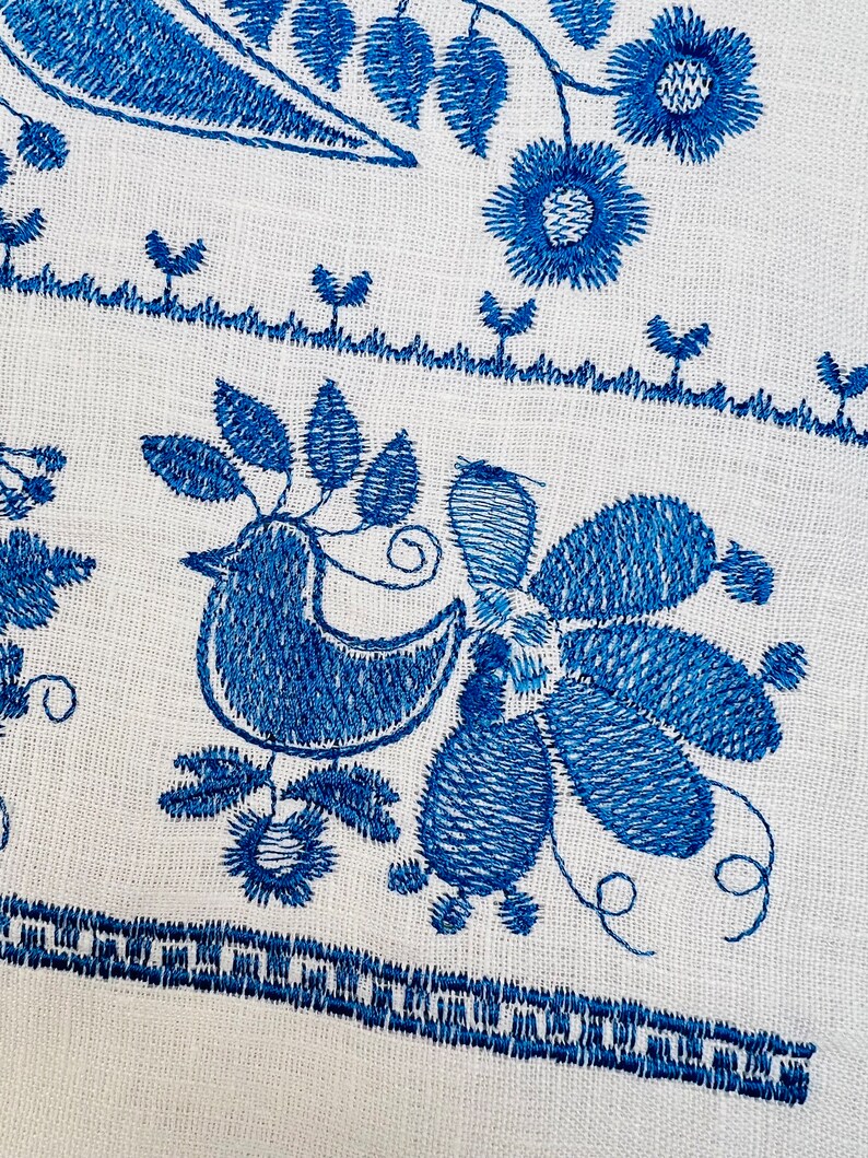 Peut inclure: Gros plan sur un tissu blanc avec une broderie bleue complexe. Le motif repr&eacute;sente des oiseaux, des fleurs et des feuilles stylis&eacute;s. La broderie est d&eacute;taill&eacute;e avec divers points, cr&eacute;ant un motif textur&eacute; et d&eacute;coratif. L'esth&eacute;tique g&eacute;n&eacute;rale est de l'art populaire.