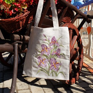 May include: A white linen tote bag with embroidered purple irises. The bag is hanging on a wooden cart with a red wheel.