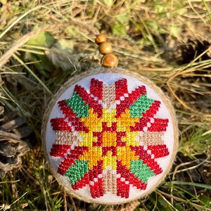 May include: A round, white, fabric ornament with a red, yellow, and green cross-stitch design. The ornament is surrounded by brown burlap and has a wooden bead string attached.