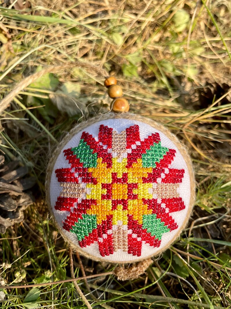 May include: A round, white, fabric ornament with a red, yellow, and green cross-stitch design. The ornament is surrounded by brown burlap and has a wooden bead string attached.