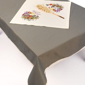 Op de afbeelding: Een grijze tafelkleed met een witte kantversiering. Een witte placemat met een geborduurd paasontwerp en de tekst "Prettige Pasen" ligt op het tafelkleed.