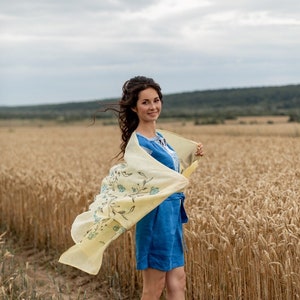 May include: A woman in a blue linen dress with a yellow floral scarf stands in a field of wheat. The scarf is blowing in the wind.