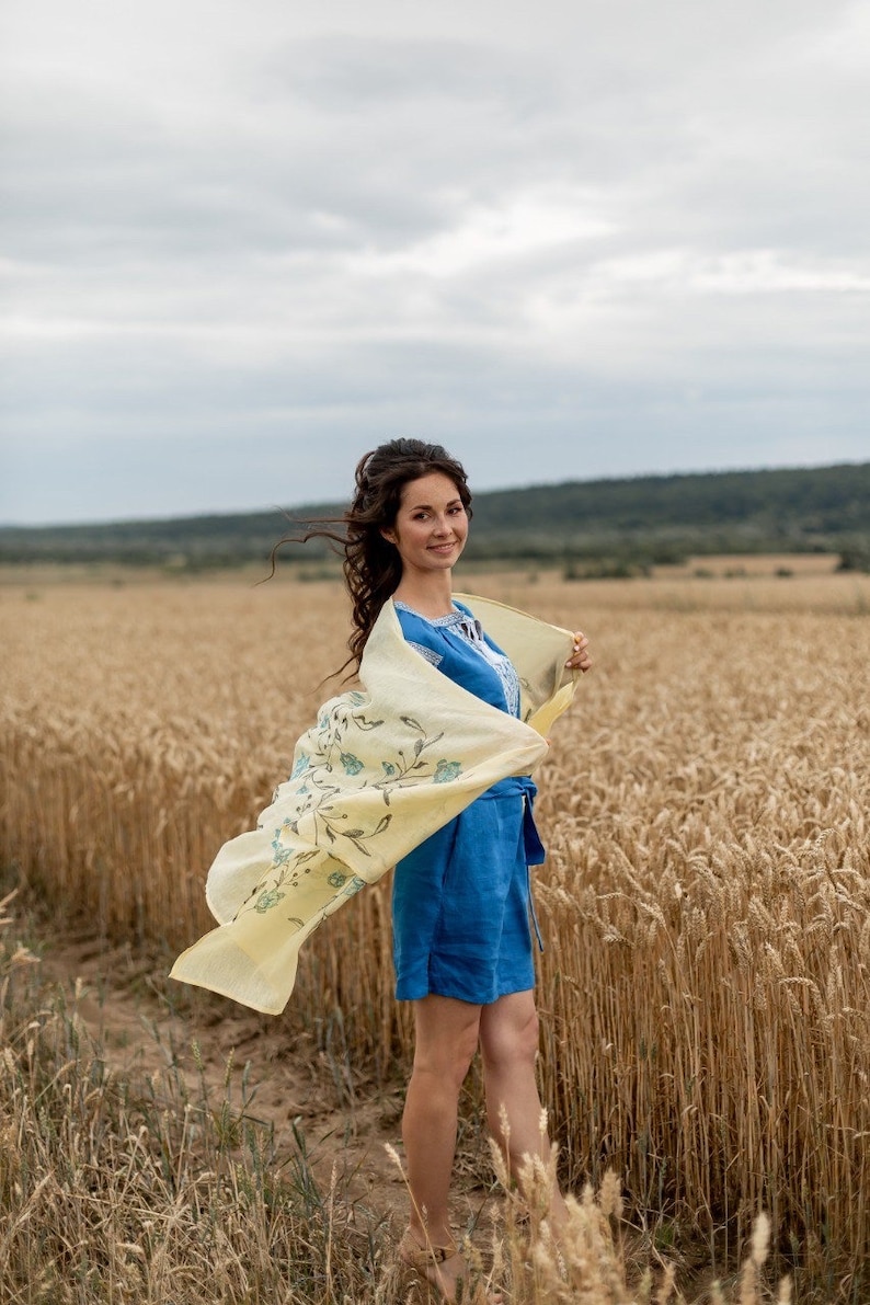 May include: A woman in a blue linen dress with a yellow floral scarf stands in a field of wheat. The scarf is blowing in the wind.