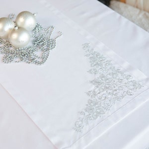 May include: White table runner with silver floral embroidery on one corner. Three silver Christmas ornaments and a silver beaded garland are arranged on the runner, creating a festive holiday display.