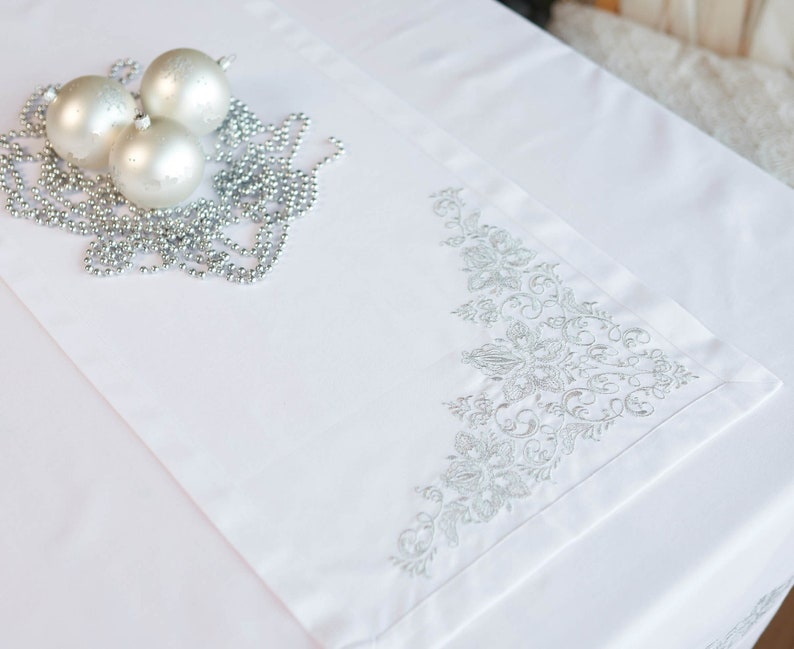 May include: White table runner with silver floral embroidery on one corner. Three silver Christmas ornaments and a silver beaded garland are arranged on the runner, creating a festive holiday display.