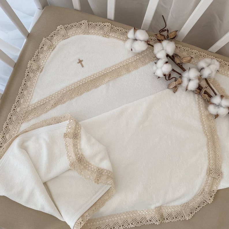 May include: White baby blanket with a cross embroidered on it. The blanket has a lace trim and is folded in half. The blanket is lying on a white crib mattress.