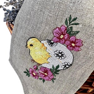 May include: A linen Easter napkin with an embroidered design. The design shows a yellow chick hatching from a white egg decorated with black flowers, surrounded by pink flowers and green leaves. The napkin is folded over a wicker basket.