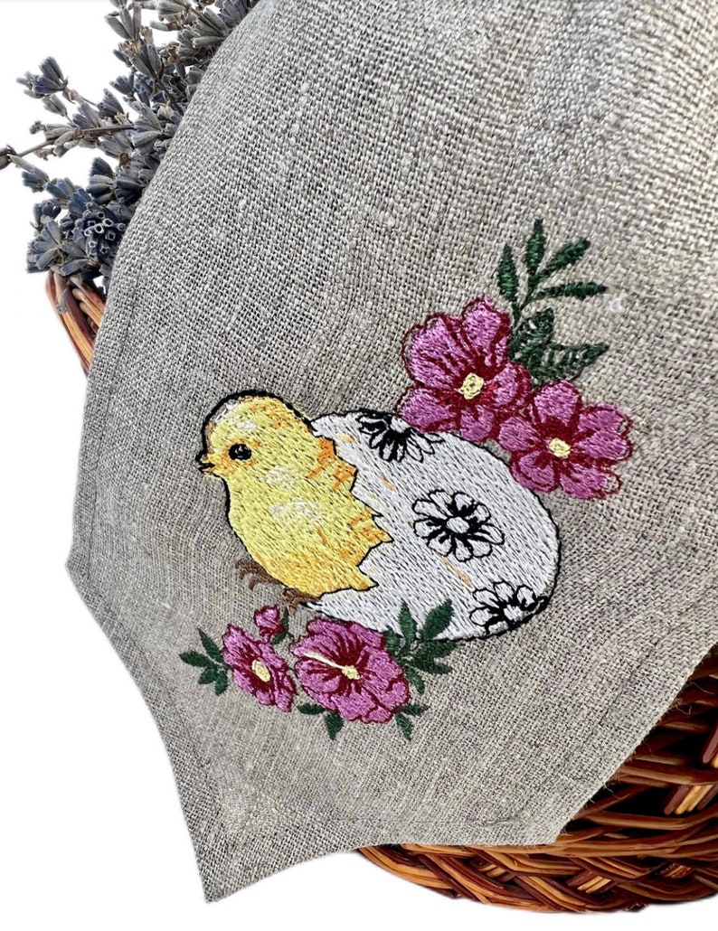 May include: A linen Easter napkin with an embroidered design. The design shows a yellow chick hatching from a white egg decorated with black flowers, surrounded by pink flowers and green leaves. The napkin is folded over a wicker basket.