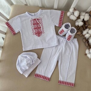 May include: A white baby outfit with red embroidered details. The outfit includes a short-sleeved shirt, pants, a hat, and booties.