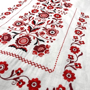 K&ouml;nnte beinhalten: Wei&szlig;er Leinenstoff mit roten und schwarzen gestickten Blumendesigns. Die Stickerei zeigt ein zentrales Blumenmotiv mit kleineren Blumendesigns an den R&auml;ndern.