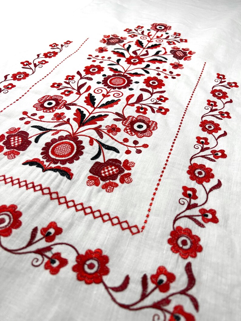 K&ouml;nnte beinhalten: Wei&szlig;er Leinenstoff mit roten und schwarzen gestickten Blumendesigns. Die Stickerei zeigt ein zentrales Blumenmotiv mit kleineren Blumendesigns an den R&auml;ndern.