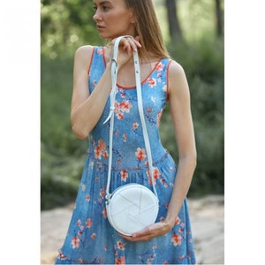 May include: A white round crossbody bag with a woven design. The bag is worn by a person wearing a blue floral dress.
