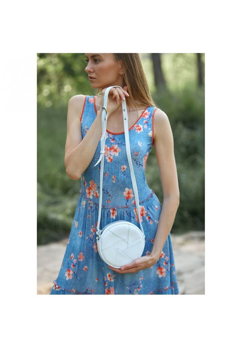 May include: A white round crossbody bag with a woven design. The bag is worn by a person wearing a blue floral dress.