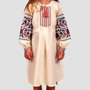 May include: A young girl wearing a white linen dress with red and blue embroidered flowers on the sleeves. The dress has a tassel detail at the neckline.
