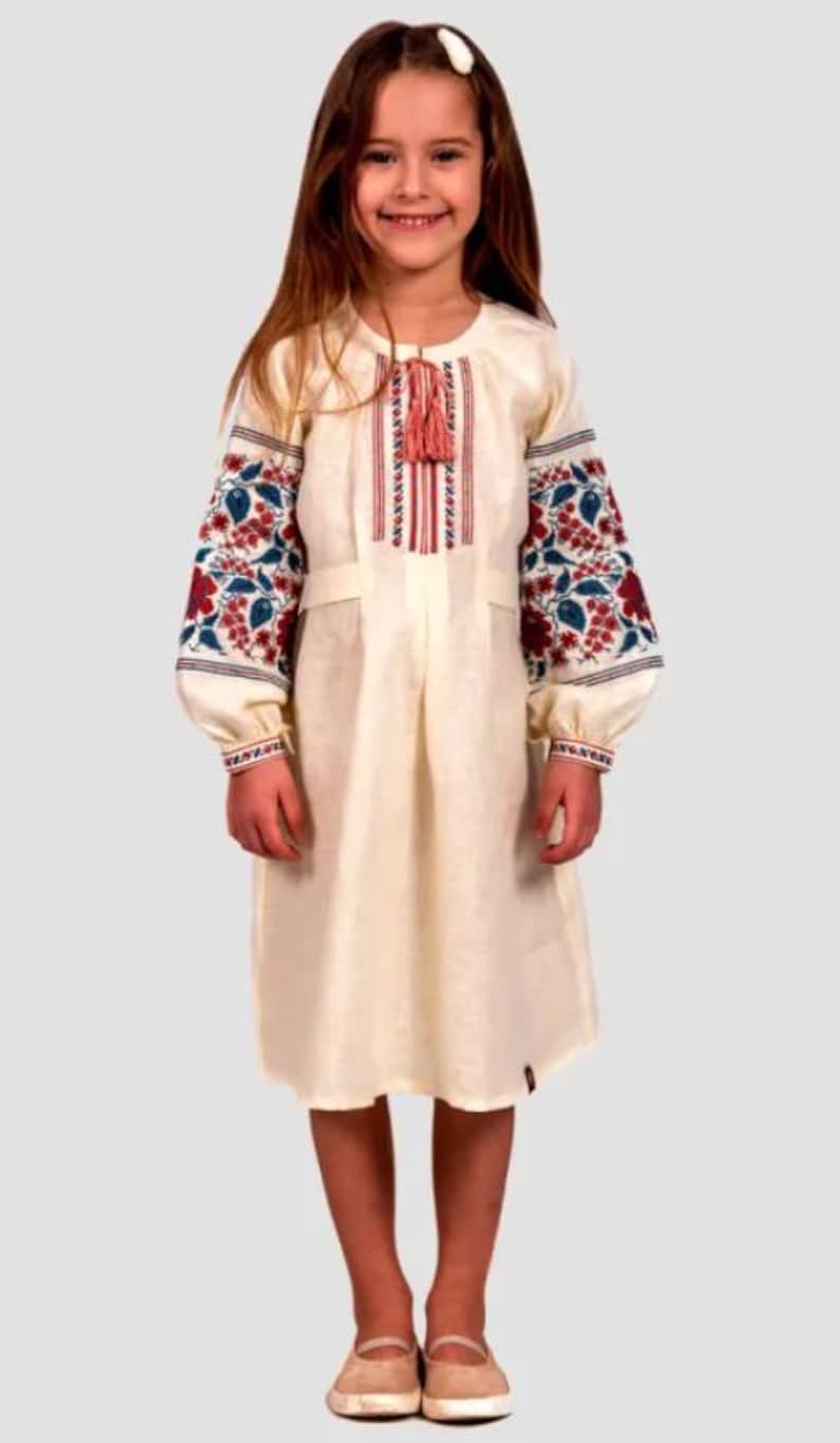 May include: A young girl wearing a white linen dress with red and blue embroidered flowers on the sleeves. The dress has a tassel detail at the neckline.