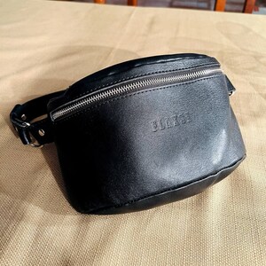 May include: A black leather fanny pack with a silver zipper and the word "BLACK" embossed on the front.