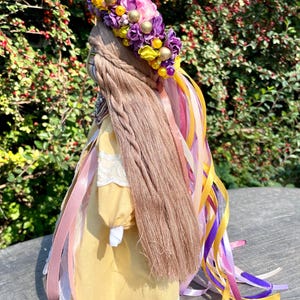 Puede incluir: Una mu&ntilde;eca de tela amarilla con cabello largo y casta&ntilde;o, que lleva un vestido de encaje blanco y una corona de flores con flores moradas y rosas y cuentas amarillas. La mu&ntilde;eca tiene varios lazos rosas, morados y amarillos atados a su cabello.