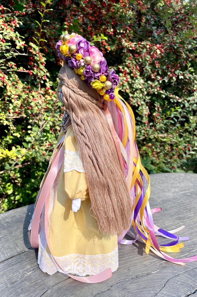 Puede incluir: Una mu&ntilde;eca de tela amarilla con cabello largo y casta&ntilde;o, que lleva un vestido de encaje blanco y una corona de flores con flores moradas y rosas y cuentas amarillas. La mu&ntilde;eca tiene varios lazos rosas, morados y amarillos atados a su cabello.