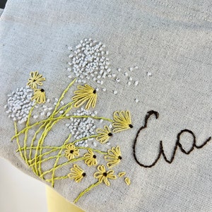May include: A light gray canvas tote bag with embroidered yellow flowers and green stems. The name "Eva" is embroidered in black cursive lettering.