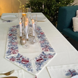 Puede incluir: Una mesa festiva con un mantel color crema, adornada con un camino de mesa decorativo con bordados de adornos navide&ntilde;os y ramas de pino. Las copas de champ&aacute;n y las velas encendidas se suman al ambiente festivo. Adornos dorados y plateados est&aacute;n colocados sobre la mesa.