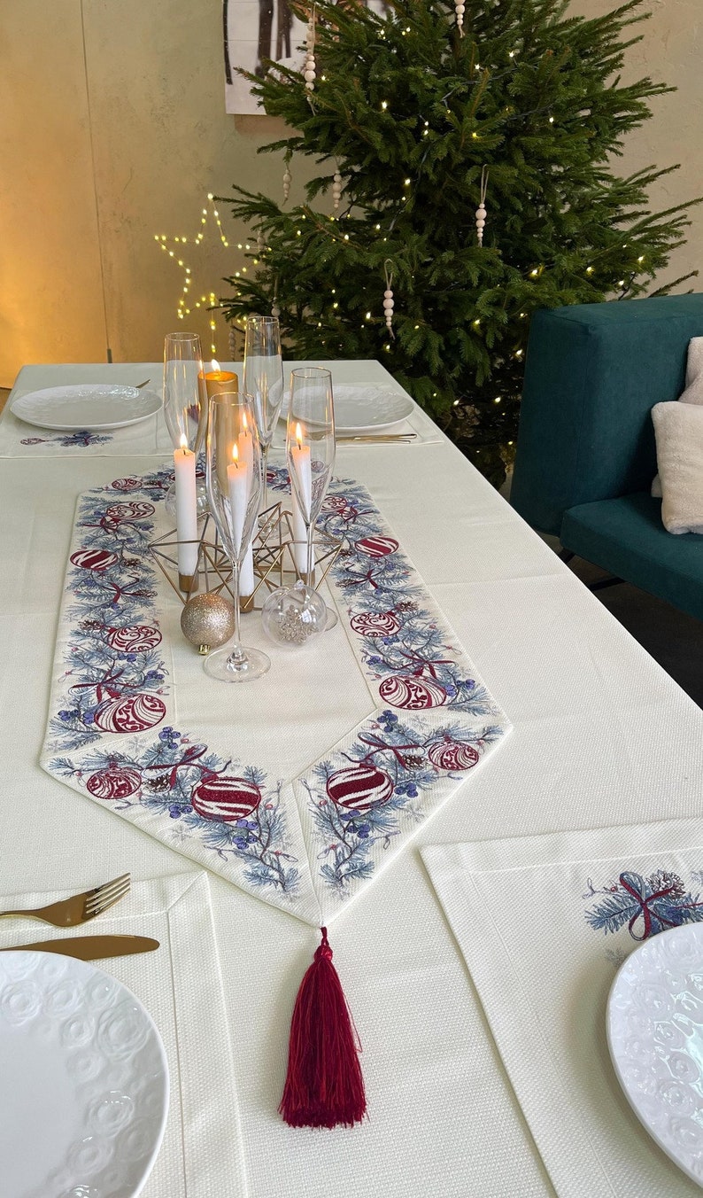 Puede incluir: Una mesa festiva con un mantel color crema, adornada con un camino de mesa decorativo con bordados de adornos navide&ntilde;os y ramas de pino. Las copas de champ&aacute;n y las velas encendidas se suman al ambiente festivo. Adornos dorados y plateados est&aacute;n colocados sobre la mesa.