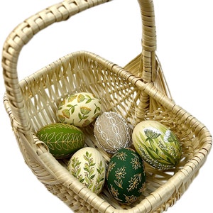 May include: A wicker basket filled with six hand-painted Easter eggs. The eggs are decorated with green and gold floral patterns.