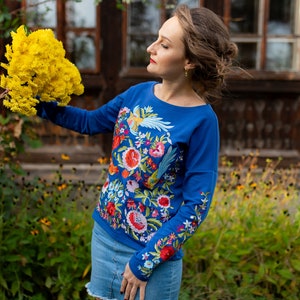Puede incluir: Una mujer con una camisa azul de manga larga con bordados florales coloridos y una falda vaquera sostiene un ramo de flores amarillas.
