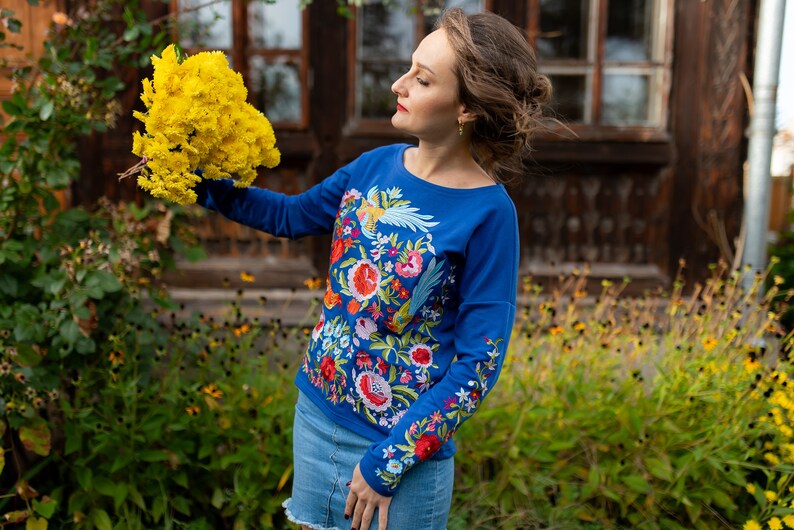 Puede incluir: Una mujer con una camisa azul de manga larga con bordados florales coloridos y una falda vaquera sostiene un ramo de flores amarillas.