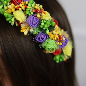 K&ouml;nnte beinhalten: Ein farbenfroher Blumenkranz mit k&uuml;nstlichen Blumen. Der Kranz enth&auml;lt rote, gelbe, lila und gr&uuml;ne Blumen mit kleinen gelben und gr&uuml;nen Akzenten. Die Blumen sind auf einer flexiblen Basis angeordnet, die zum Tragen geeignet ist.
