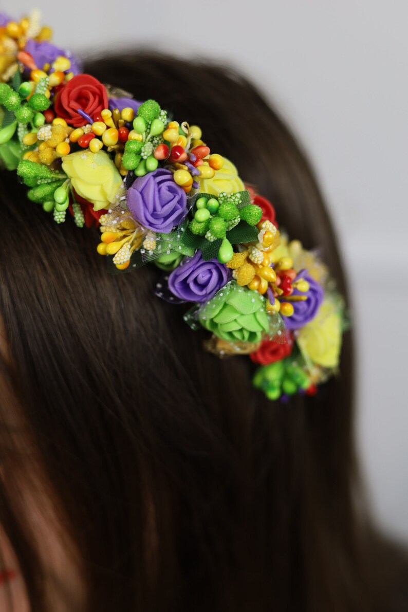 K&ouml;nnte beinhalten: Ein farbenfroher Blumenkranz mit k&uuml;nstlichen Blumen. Der Kranz enth&auml;lt rote, gelbe, lila und gr&uuml;ne Blumen mit kleinen gelben und gr&uuml;nen Akzenten. Die Blumen sind auf einer flexiblen Basis angeordnet, die zum Tragen geeignet ist.