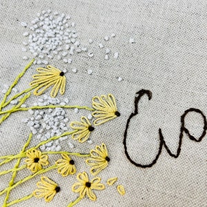 May include: Embroidered design on a beige fabric featuring a cluster of yellow flowers with black centers and a white embroidered name "Eva" in cursive.