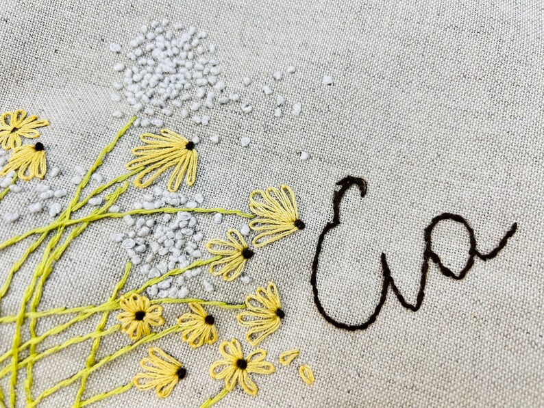 May include: Embroidered design on a beige fabric featuring a cluster of yellow flowers with black centers and a white embroidered name "Eva" in cursive.