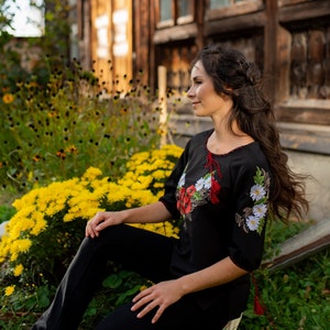 May include: A woman wearing a black embroidered blouse with red and white floral designs. She is sitting in front of a yellow flower bush and looking to the right.