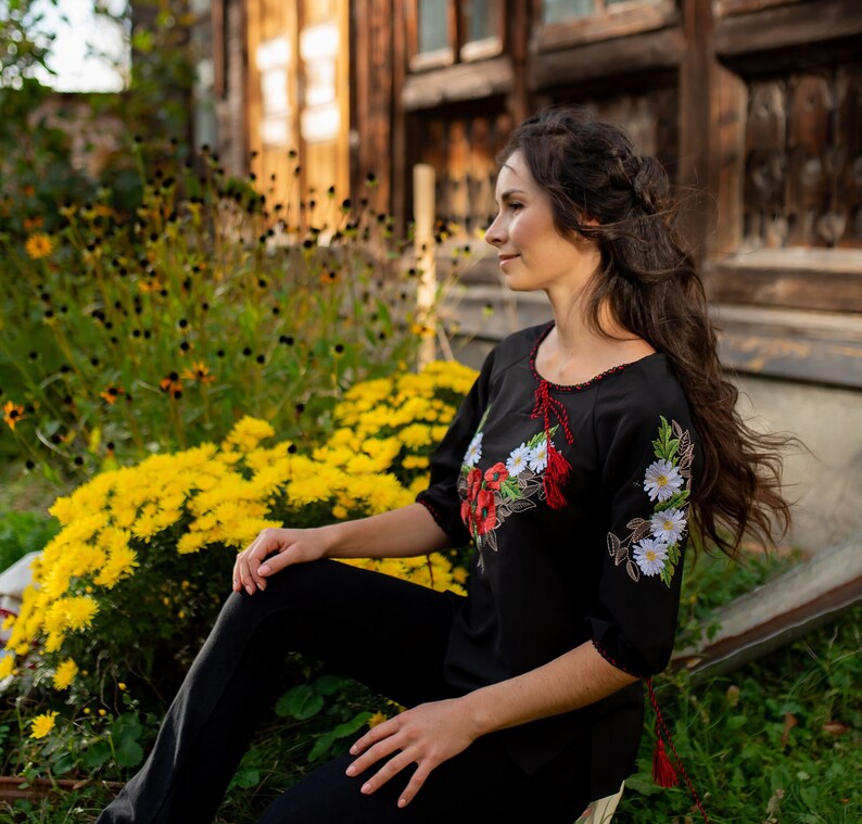 May include: A woman wearing a black embroidered blouse with red and white floral designs. She is sitting in front of a yellow flower bush and looking to the right.