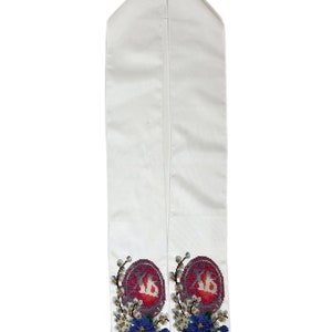 May include: White graduation stole with embroidered details. Features a maroon emblem with white lettering, surrounded by blue and white floral embroidery. The stole is made of a smooth, satin-like material.
